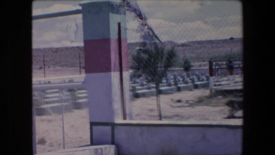 1971 Algeria 8mm frame with a red-and-white wall post in the foreground and a fenced memorial enclosure beyond, showing rows of markers, sparse plants, and arid open land.
