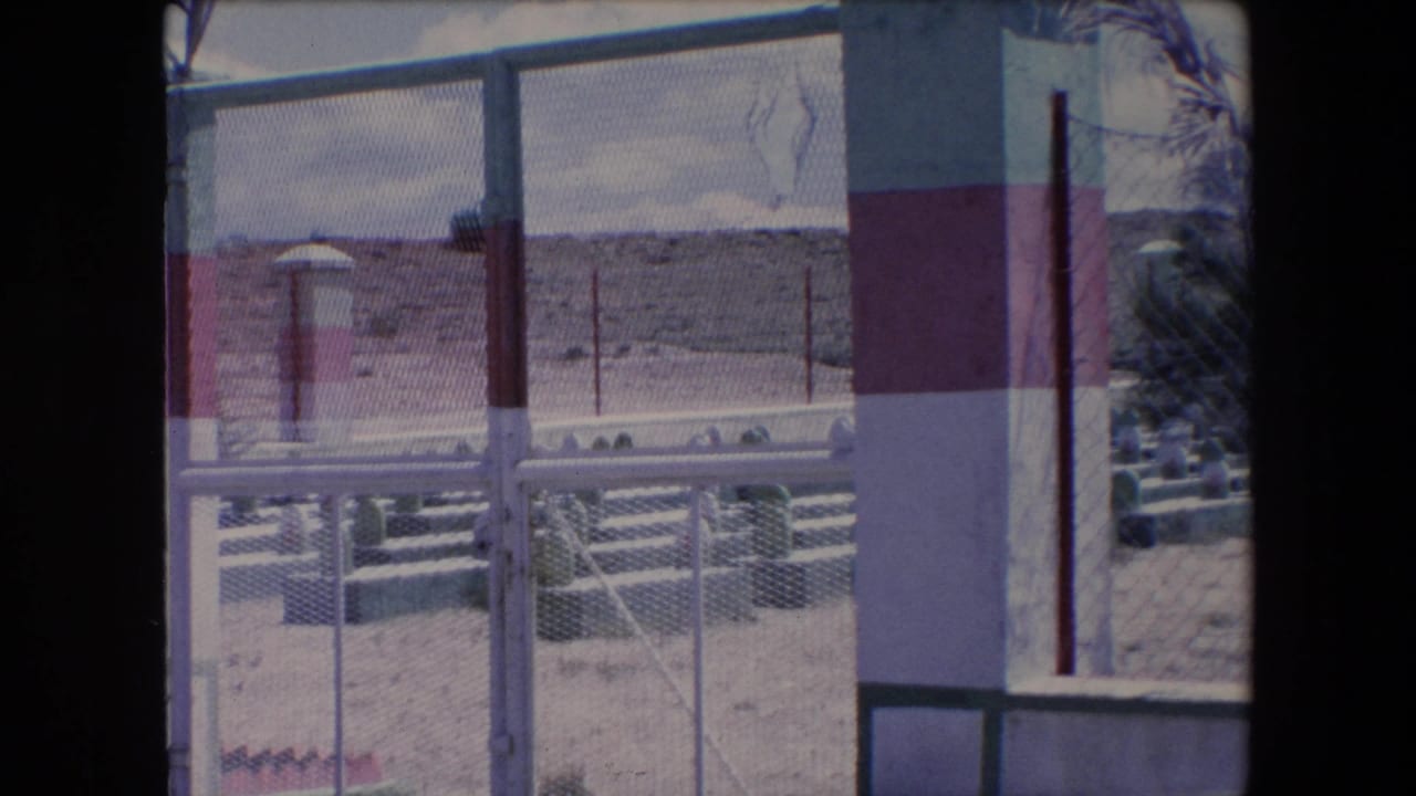 1971 Algeria home-movie frame with a chain-link fence and striped masonry pillar in front of a sandy cemetery, white grave markers, sparse plants, and low hills beyond.