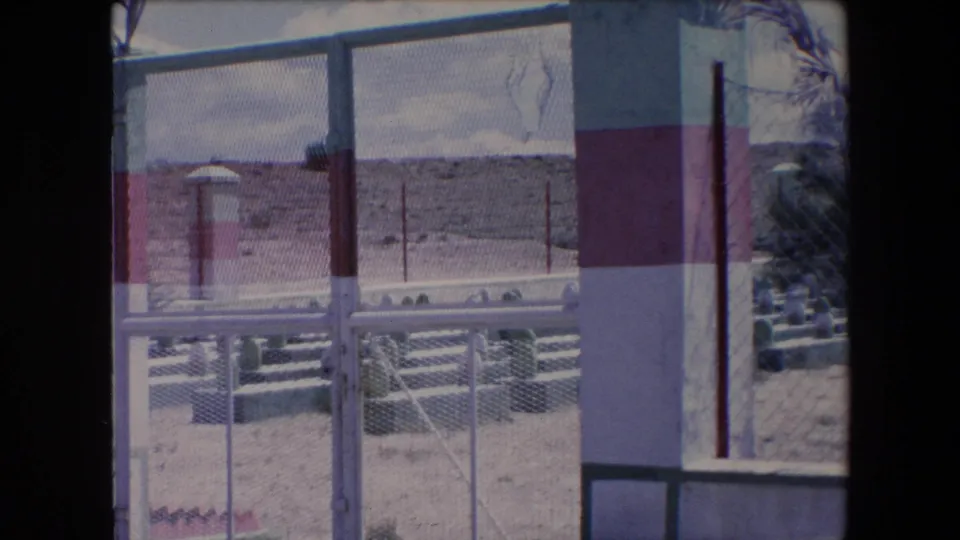 1971 Algeria home-movie frame with a chain-link fence and striped masonry pillar in front of a sandy cemetery, white grave markers, sparse plants, and low hills beyond.