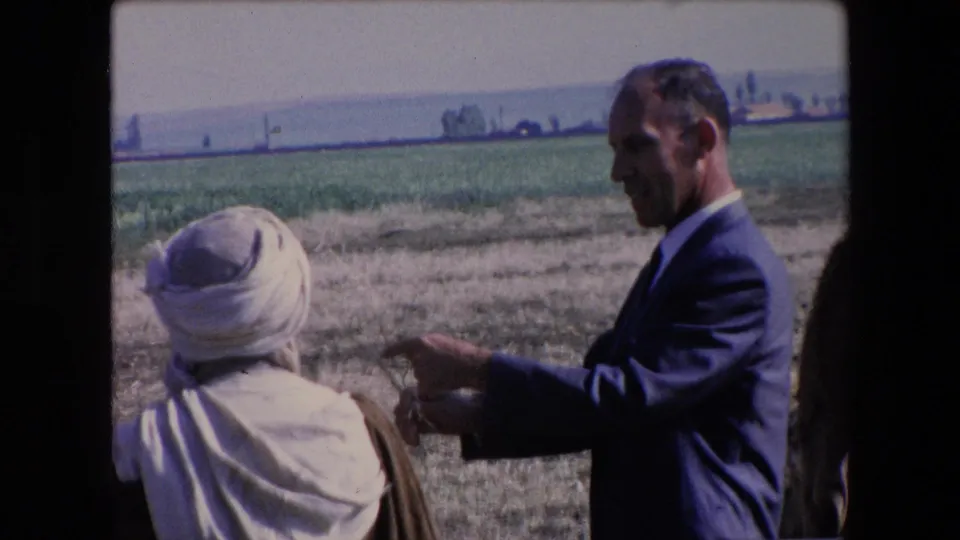 1971 Algeria 8mm frame showing a suited adult man pointing near a person with a white head covering, with camels and fields around the dry pasture.
