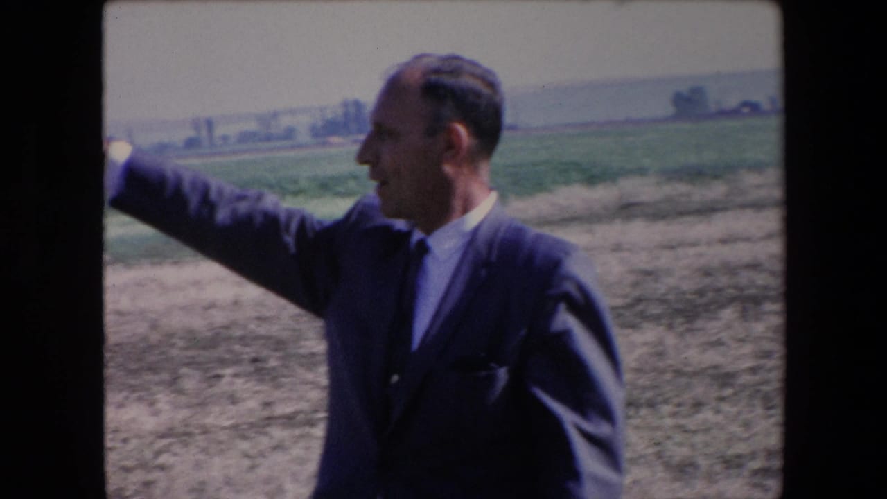 A suited man in 1971 Algeria raises his arm in a broad gesture beside dry ground and green agricultural fields under bright daylight.
