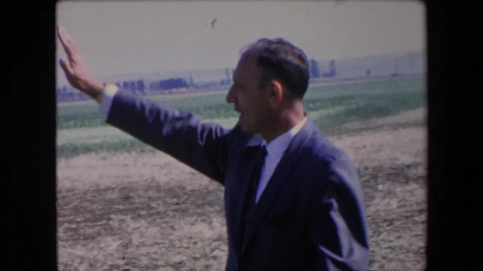 The suited man continues gesturing in the Algeria field scene, with his profile visible against dry ground, green crops, and distant trees.