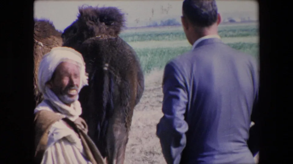 1971 Algeria 8mm frame with a smiling man in traditional white headwear looking toward the camera, a camel at center, and a suited traveler seen from behind.