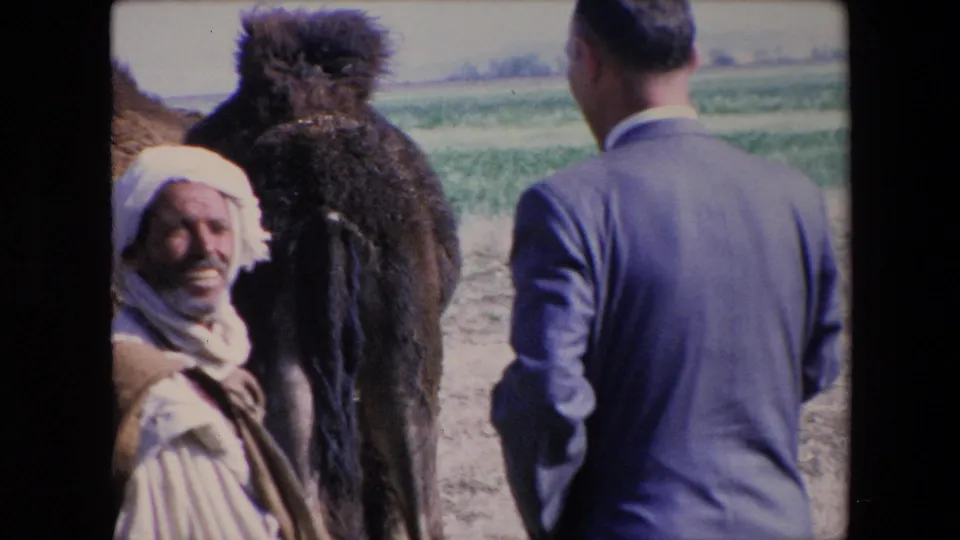 1971 Algeria 8mm frame showing an older man in white clothing and headwrap smiling near a camel while a suited visitor stands with his back to camera.