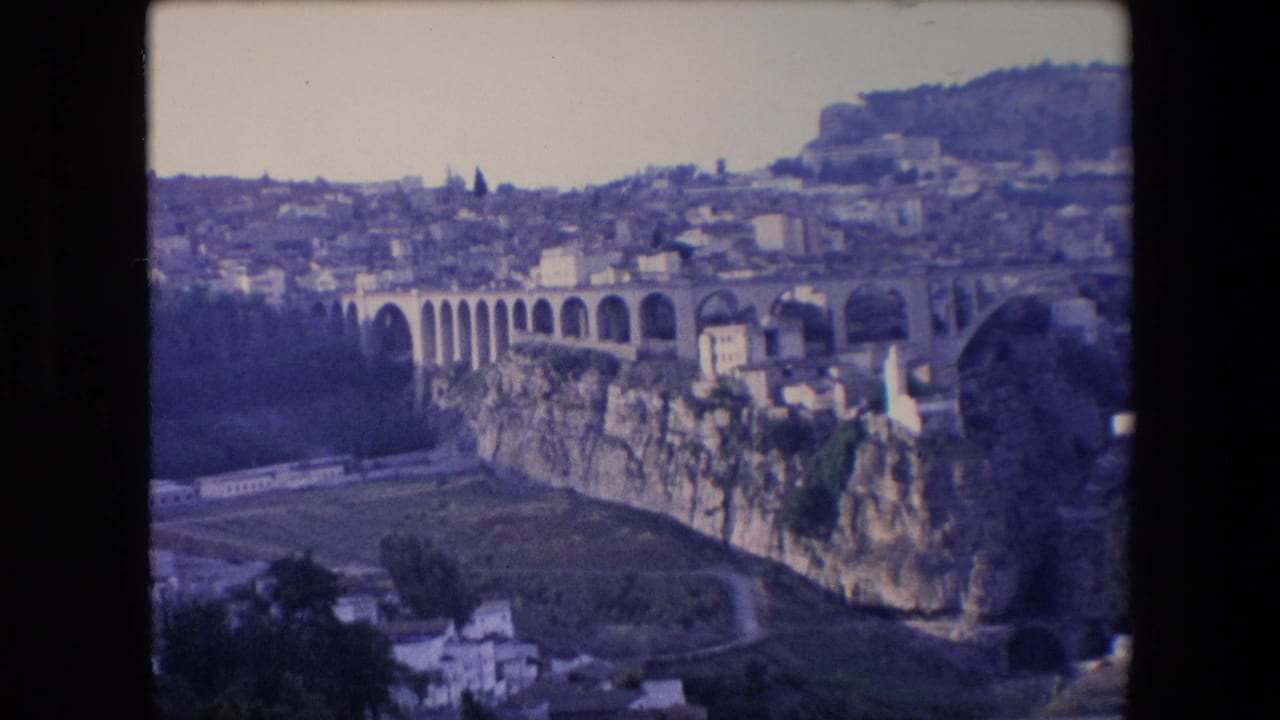 1971 Algeria 8mm frame with a wide visitor-shot view of a large arched stone aqueduct-like structure crossing a deep ravine below clustered hillside buildings.