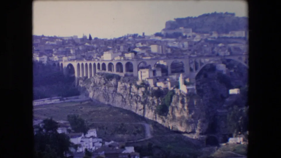 1971 Algeria 8mm frame continuing the panoramic overlook of arched stonework above a rocky ravine, with pale buildings spread across the hillside beyond.