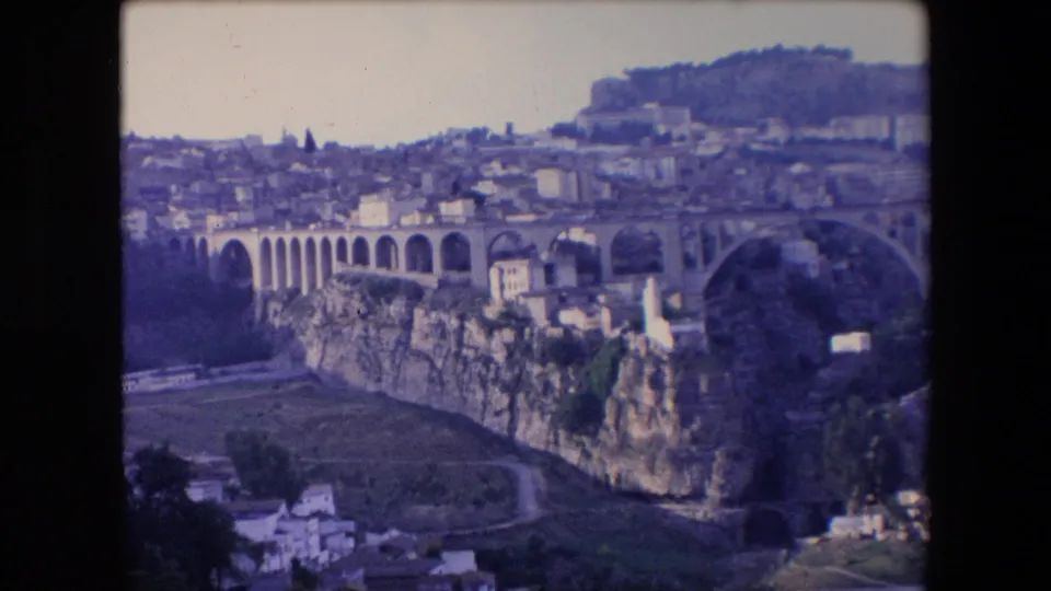 1971 Algeria 8mm frame showing the aqueduct-like arches and rocky ravine from a scenic overlook, with a hillside settlement and plateau in the background.
