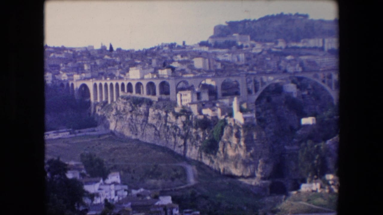 1971 Algeria 8mm frame showing a large arched bridge over a rocky ravine, with clustered white buildings and hillside structures under a pale sky.
