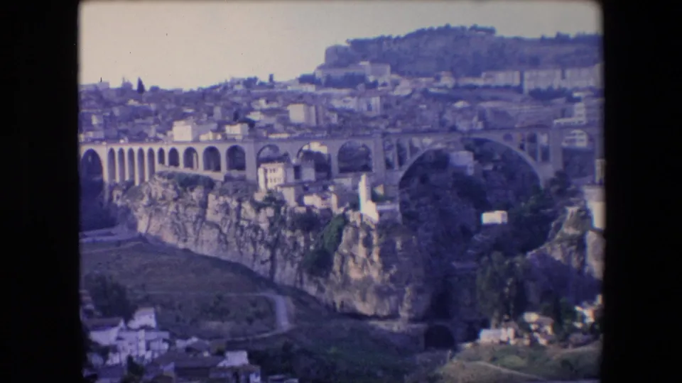 1971 Algeria 8mm frame with a wide view of an arched bridge, steep rocky cliffs, hillside buildings, and a pale sky in faded color.