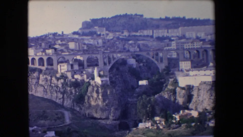 1971 Algeria 8mm frame showing the bridge arch more prominently, with rocky ravine walls, hillside structures, and small white buildings below.