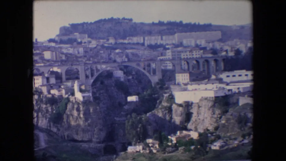 1971 Algeria 8mm frame showing the arched bridge over a deep ravine, with white buildings, hillside development, and a bright road or structure at right.