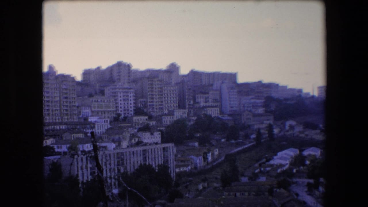 1971 Algeria home-movie frame looking over a dense hillside cityscape with mid-rise apartment blocks, smaller homes, trees, and dark 8mm film borders.