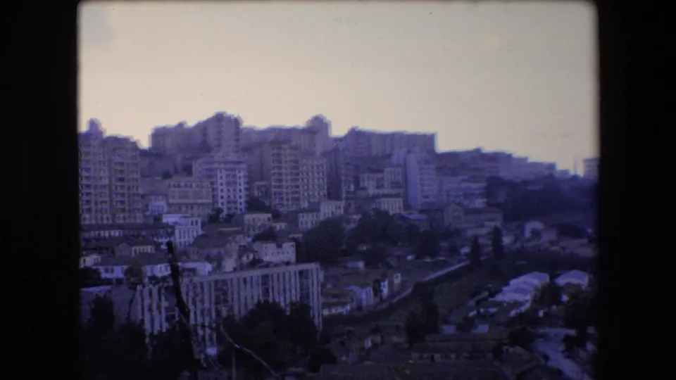 1971 Algeria home-movie frame looking over a dense hillside cityscape with mid-rise apartment blocks, smaller homes, trees, and dark 8mm film borders.