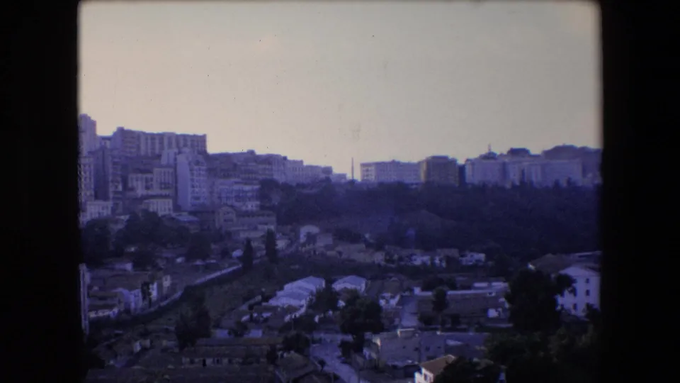 1971 Algeria home-movie still holding on a hillside urban district with apartment blocks, smaller buildings, trees, and the visible edges of the 8mm scan.