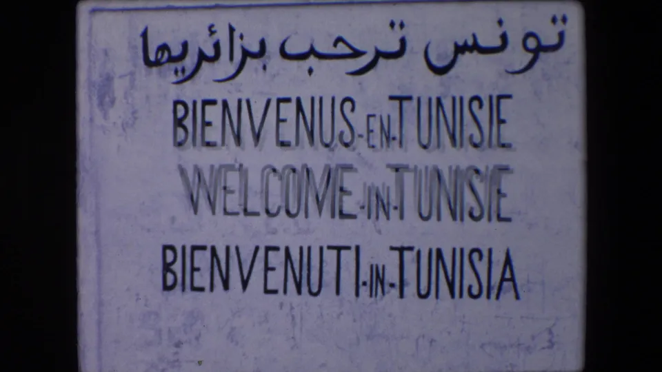 Roadside sign in the 1971 travel footage reads as a multilingual welcome to Tunisia, with Arabic script above French, English, and Italian lines.