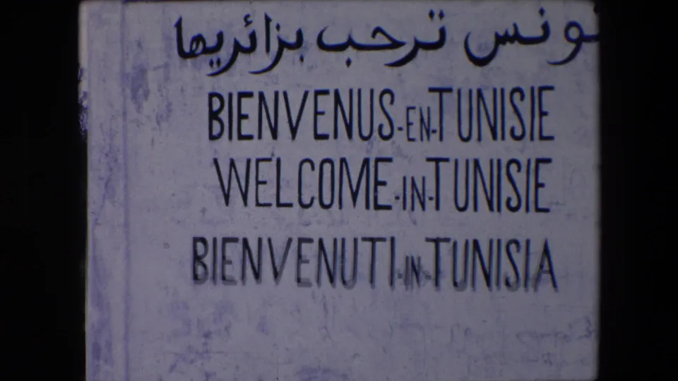 The frame centers on a weathered multilingual welcome sign for Tunisia, showing Arabic script and French, English, and Italian wording in aged 8mm color.
