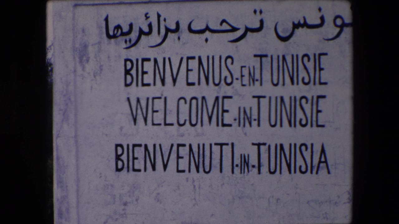 The 8mm frame holds on the Tunisie welcome sign, with Arabic script and French, English, and Italian welcome lines visible on a worn pale surface.