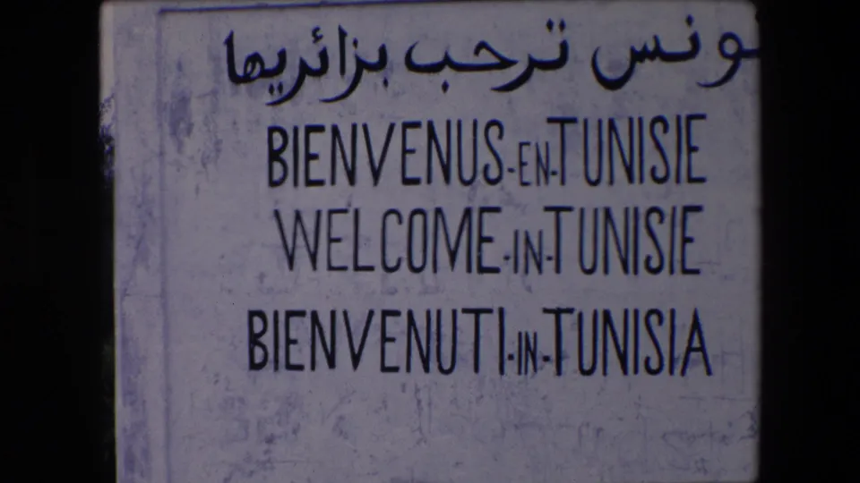 The 8mm frame holds on the Tunisie welcome sign, with Arabic script and French, English, and Italian welcome lines visible on a worn pale surface.