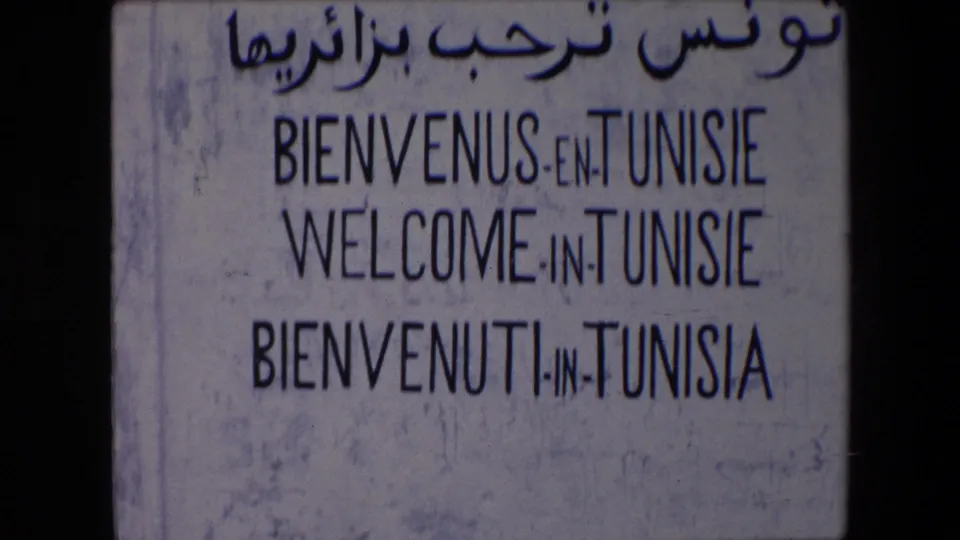 A slightly lower 1971 8mm view repeats the weathered Tunisie welcome sign, emphasizing the painted multilingual lettering and aged surface.
