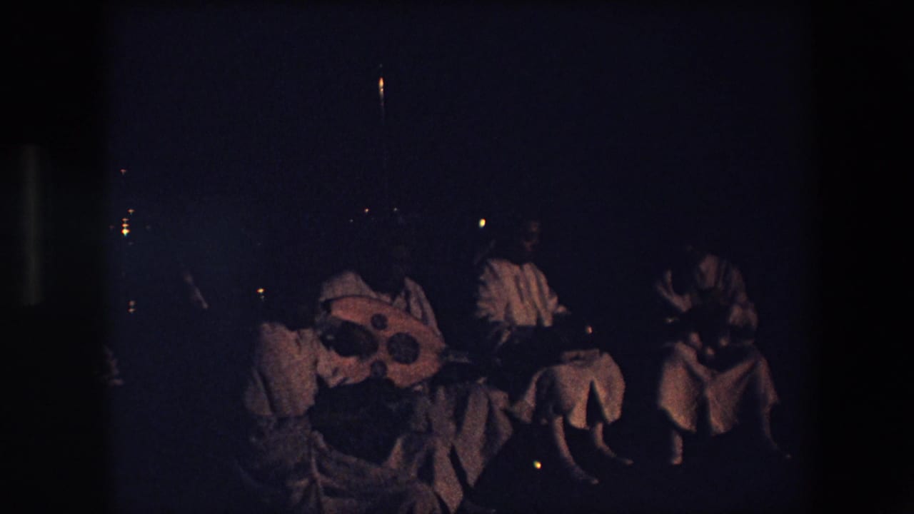 In Algeria in 1971, a dim 8mm frame shows seated adults in pale garments gathered at night with a large round lute-like instrument and small percussion nearby.