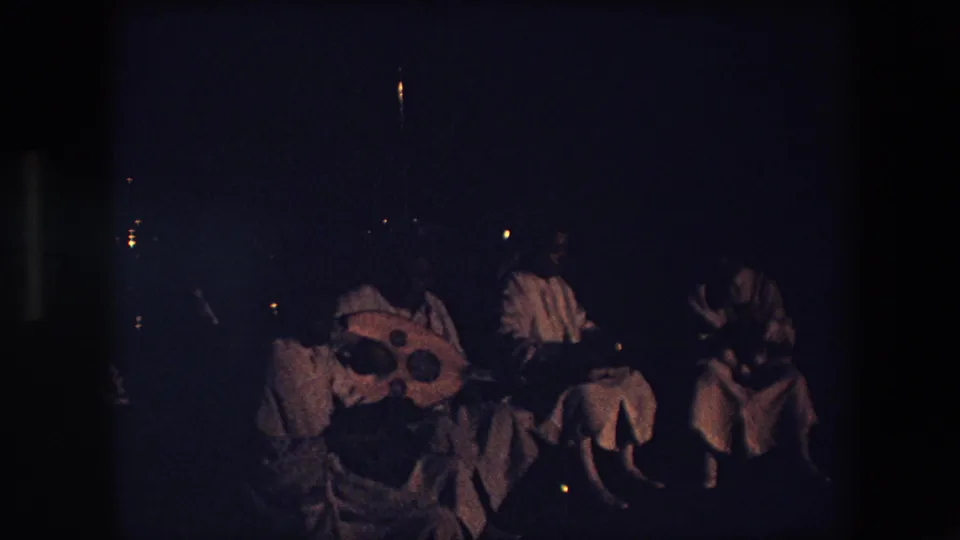 In Algeria in 1971, a dim 8mm frame shows seated adults in pale garments gathered at night with a large round lute-like instrument and small percussion nearby.