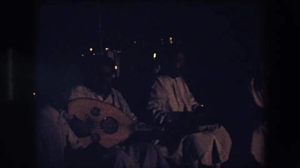 This Algeria 1971 8mm frame moves closer to the seated gathering, with the large round stringed instrument more prominent and figures partly obscured by darkness.