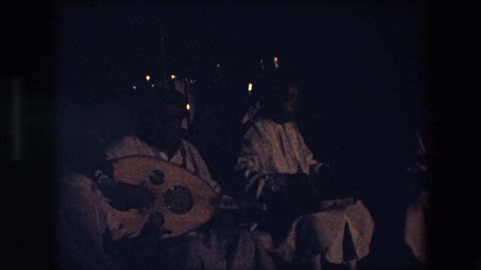 At the end of the clip, the 1971 Algeria home-movie frame shows the seated nighttime music group with pale garments, the round lute-like instrument, and surrounding darkness.
