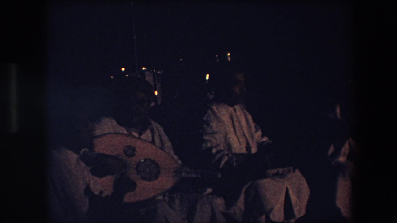 In this 1971 Algeria home-movie frame, a seated performer in light garments appears to play a rounded stringed instrument in a very dark performance space.