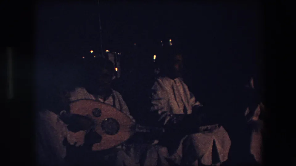 In this 1971 Algeria home-movie frame, a seated performer in light garments appears to play a rounded stringed instrument in a very dark performance space.