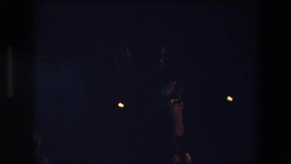 A dim 1971 Algeria home-movie frame captures a dancer in ornate costume raising an arm, with the rest of the performance area falling into deep shadow.