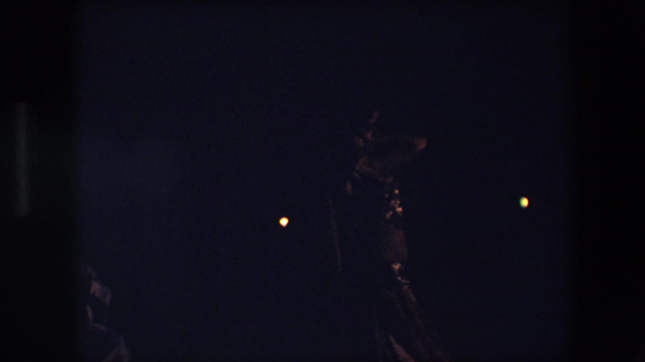 Low-light 1971 Algeria 8mm frame showing a woman in a shimmering dance outfit moving against a dark night background with scattered warm lights.