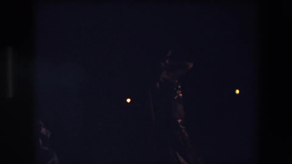 Low-light 1971 Algeria 8mm frame showing a woman in a shimmering dance outfit moving against a dark night background with scattered warm lights.