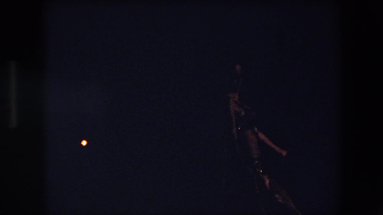 Underexposed 1971 Algeria home-movie frame with the dancer seen at the right edge, her reflective costume catching light in the dark setting.