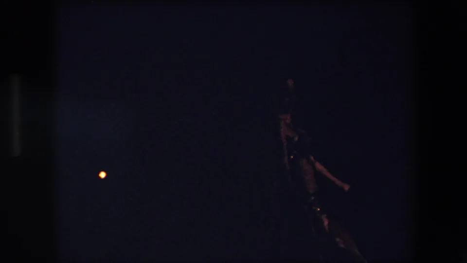 Underexposed 1971 Algeria home-movie frame with the dancer seen at the right edge, her reflective costume catching light in the dark setting.