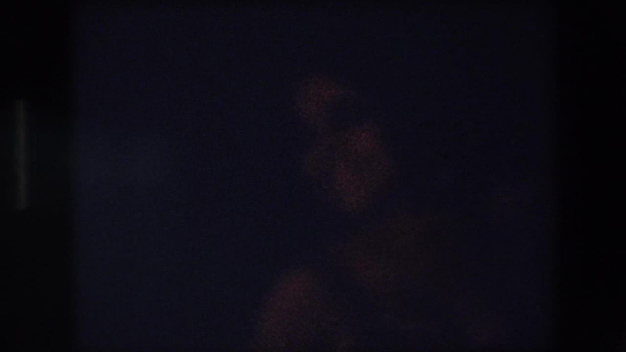 Very dark blurred frame with faint warm shapes and film grain.