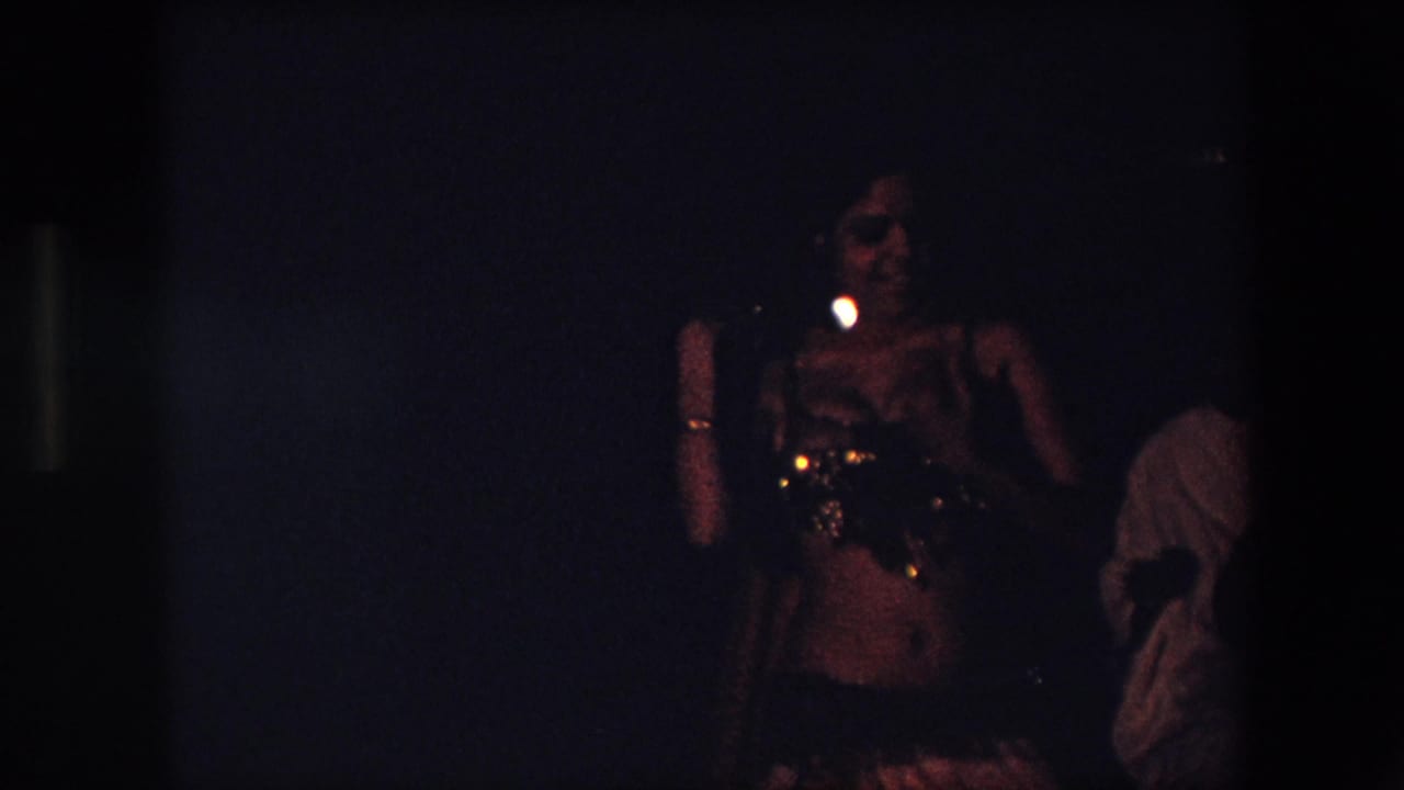 In 1971 Algeria, a belly dancer in an embellished costume performs in very dim indoor light while other people stand or sit in the background.