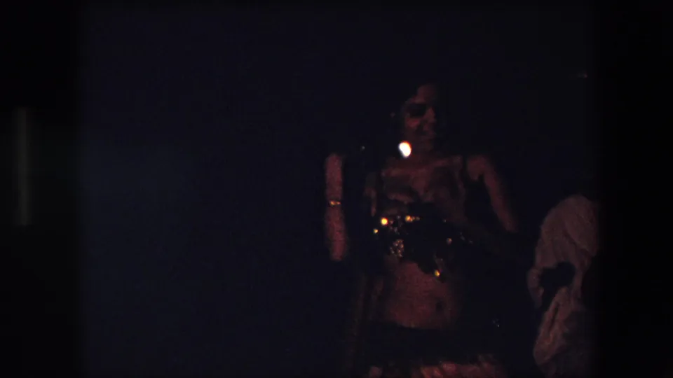 In 1971 Algeria, a belly dancer in an embellished costume performs in very dim indoor light while other people stand or sit in the background.