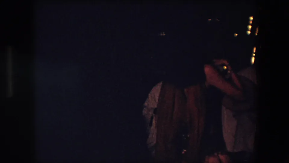 A dim 1971 Algeria frame shows the dancer partly hidden by darkness and a nearby person at the right edge of the indoor performance space.