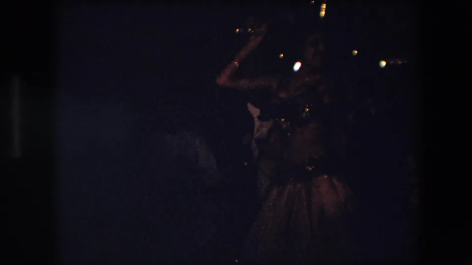 The belly dancer raises one arm during the performance, with the underexposed 8mm frame showing only faint background figures and small points of light.