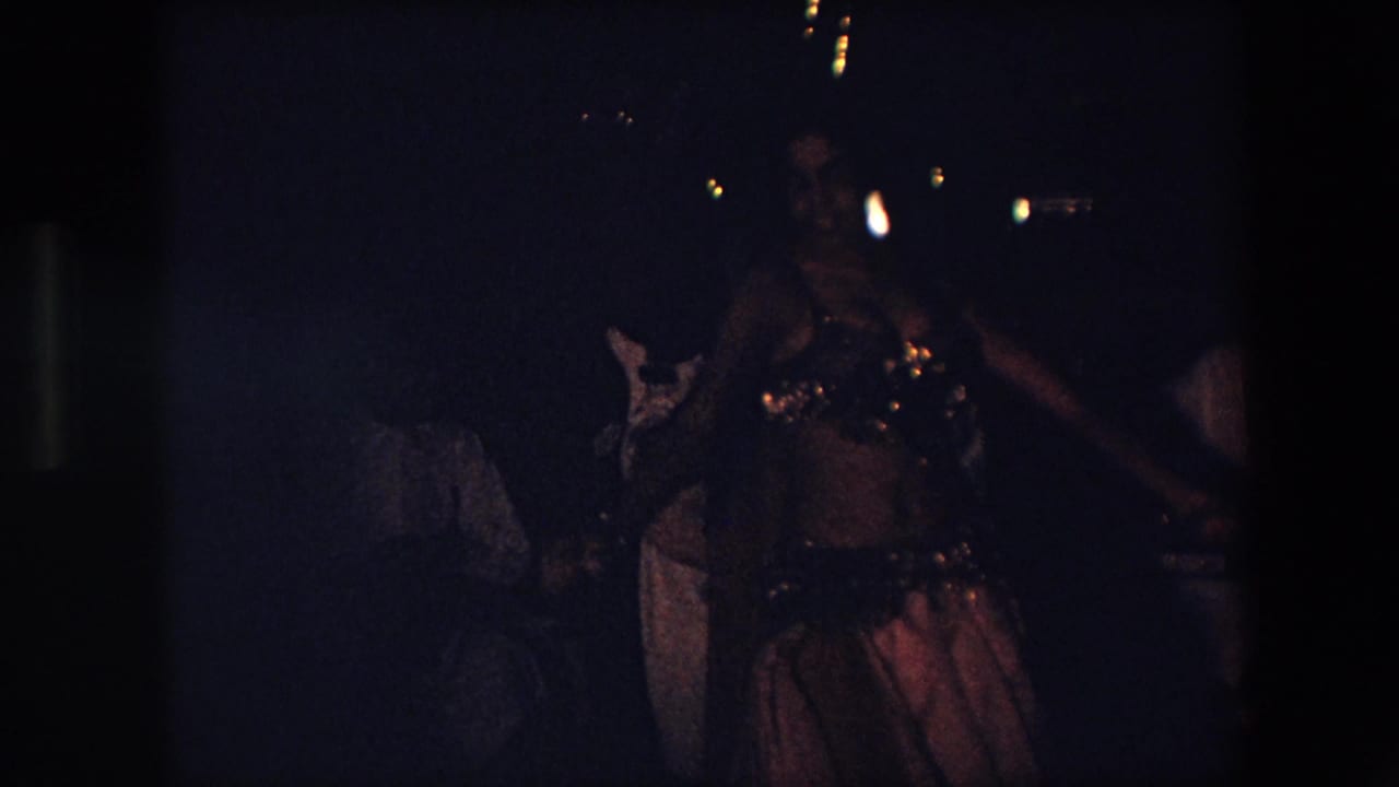 The dancer continues performing in low light, with her beaded top catching small highlights and indistinct attendees visible behind her in the Algeria 1971 footage.