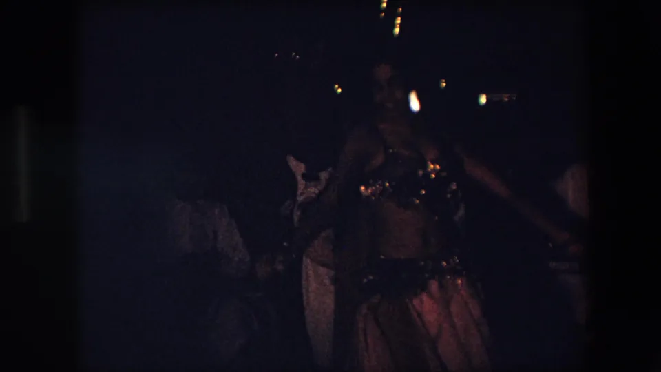 The dancer continues performing in low light, with her beaded top catching small highlights and indistinct attendees visible behind her in the Algeria 1971 footage.
