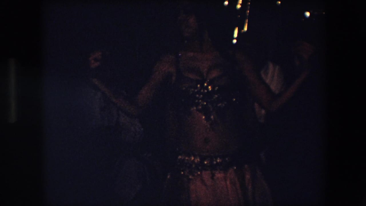 A frontal view shows the belly dancer with both arms lifted, her costume reflecting small points of light amid heavy grain and underexposed shadows.