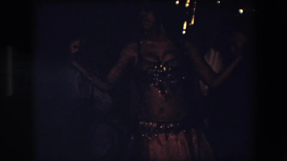A frontal view shows the belly dancer with both arms lifted, her costume reflecting small points of light amid heavy grain and underexposed shadows.