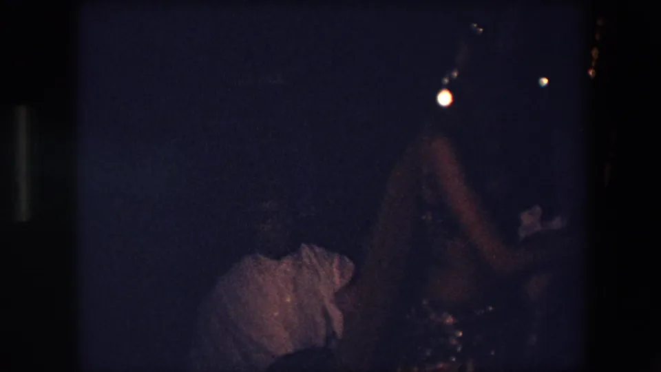 A close, underexposed 8mm frame shows the sequined dancer moving in a dark Algeria 1971 nighttime performance scene, with another figure partly visible at the lower left.