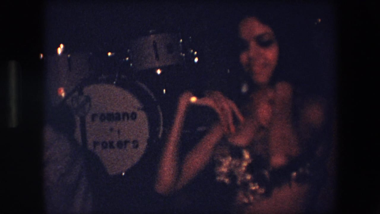 In Algeria in 1971, a belly dancer in a sparkling costume performs in dim light beside a drum kit with visible lettering on the bass drum.