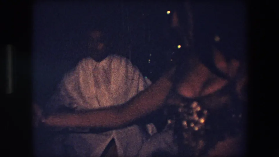 A belly dancer in sequins is partly visible beside another person in a white shirt during a dimly lit 1971 Algeria performance scene.