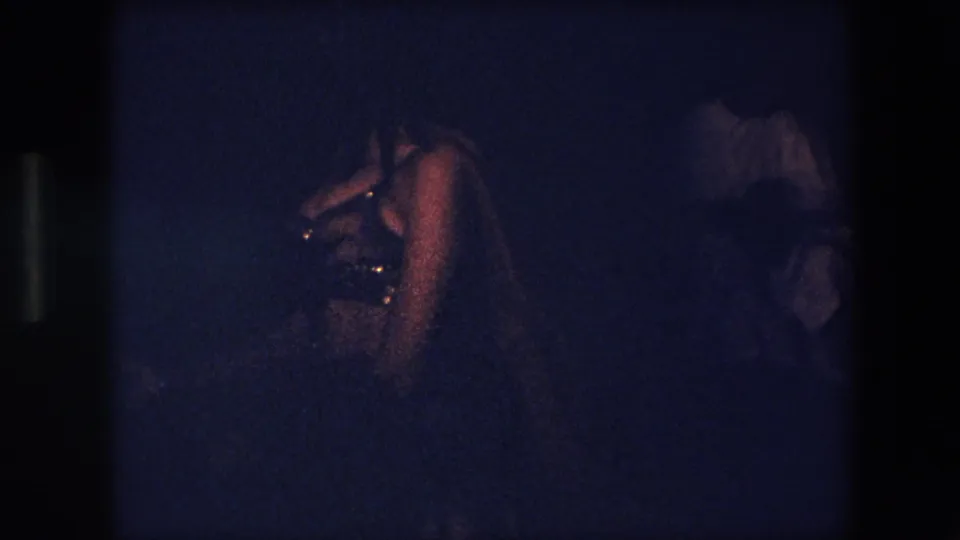 The dancer appears smaller in frame in a dim Algeria 1971 performance space, with another person partially visible at the edge of the scene.