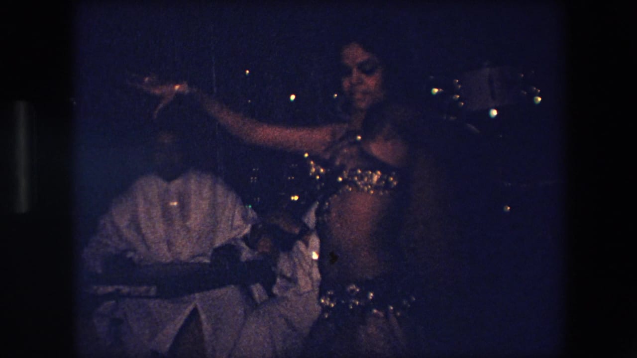 Dancer extends one arm while a seated band member is visible behind her.