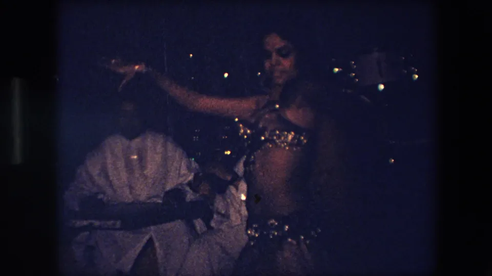 A dancer in a shimmering costume gestures with one arm in front of a seated musician in this 1971 Algeria home-movie frame.