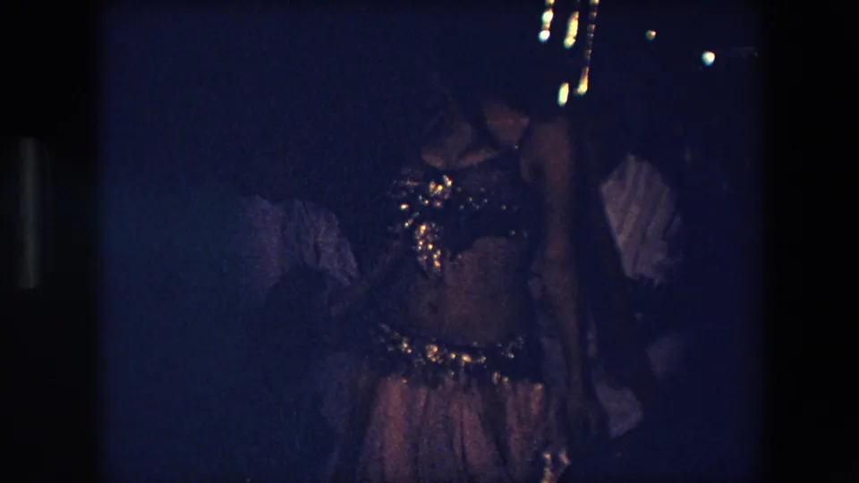 A belly dancer in a glittering costume is seen from the side near musicians, with heavy grain and dim exposure in 1971 Algeria footage.